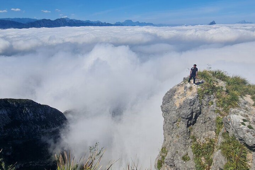 A person standing on a cliff edge overlooking a sea of clouds with mountains in the distance. 1