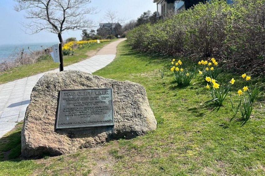 A stone sign with text, a pathway, yellow daffodils, and a grassy area. 1