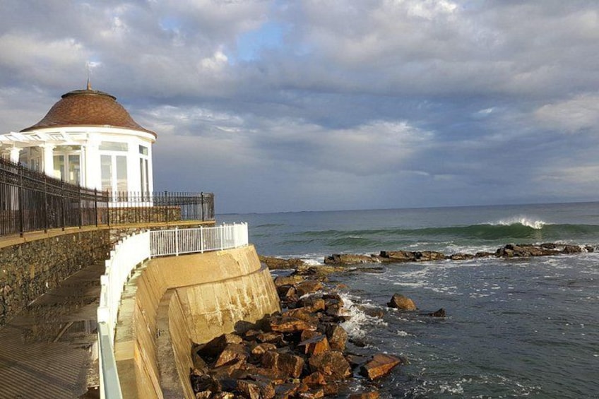 A coastal walkway with a white gazebo, rocky shoreline, and waves crashing against the rocks. 3