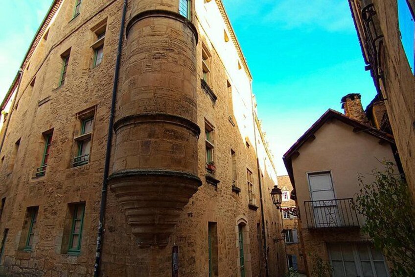 A narrow street with historic buildings, a prominent stone tower, and a small balcony. 2