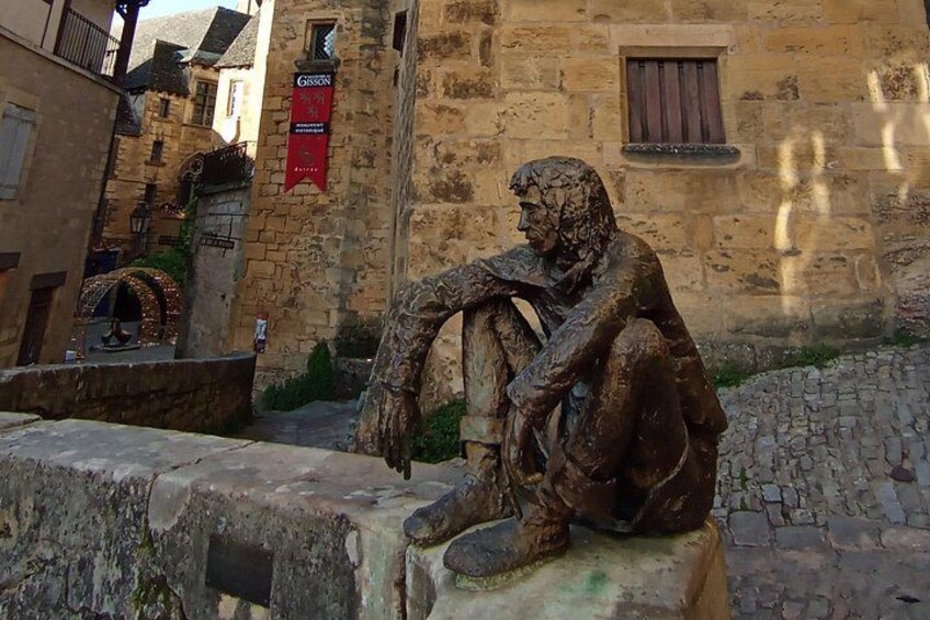 A bronze statue of a seated man in a historical setting with stone buildings and a cobblestone path. 4