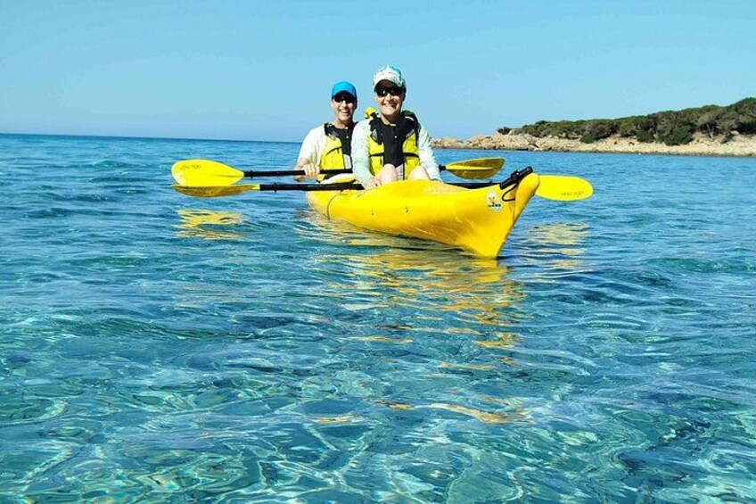 Two people in a yellow kayak on clear blue water with a beach and vegetation in the background. 2