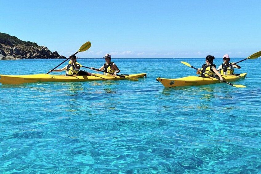 Two people kayaking on clear blue water with a rocky coastline in the background. 1