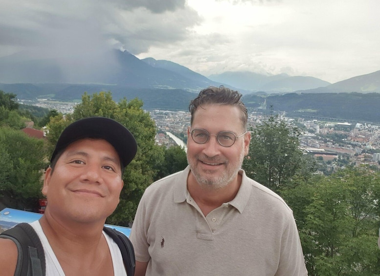 Two people standing with a city and mountains in the background. 1