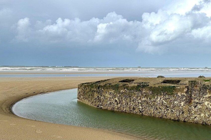 A beach with a curved stone wall, calm water, and a cloudy sky. 2