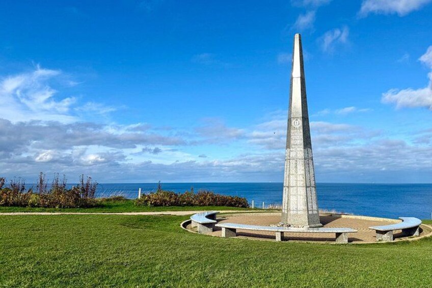A tall, slender monument with a circular base, surrounded by a grassy area and a clear blue sky. 3