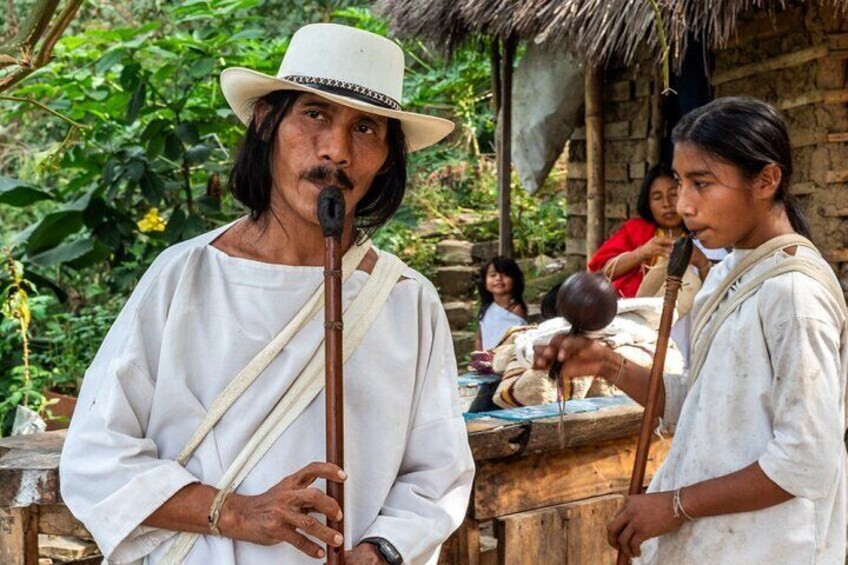 En person i traditionell dräkt som håller ett musikinstrument och står utomhus med en rustik bakgrund. 1