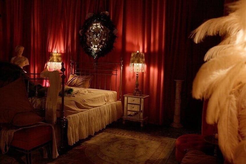 Une pièce faiblement éclairée, avec un lit, deux lampes, un rideau rouge et un fauteuil à motifs de plumes. 3