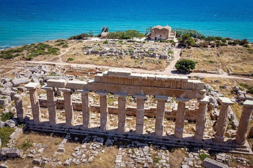 Ancient ruins with columns, a stone building, and a coastal background. 2