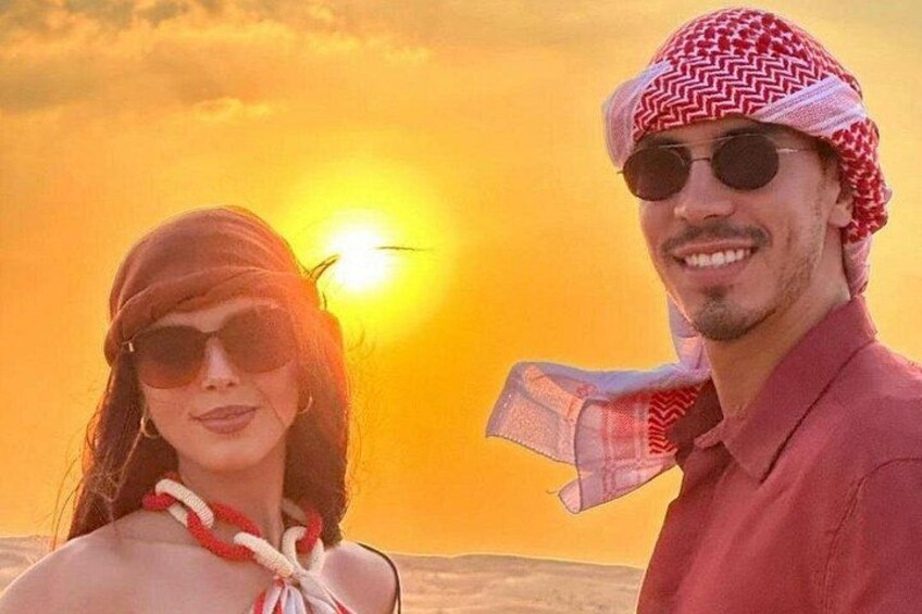 A person wearing a red shirt and a patterned headscarf, standing in a desert landscape with a sunset in the background. 4
