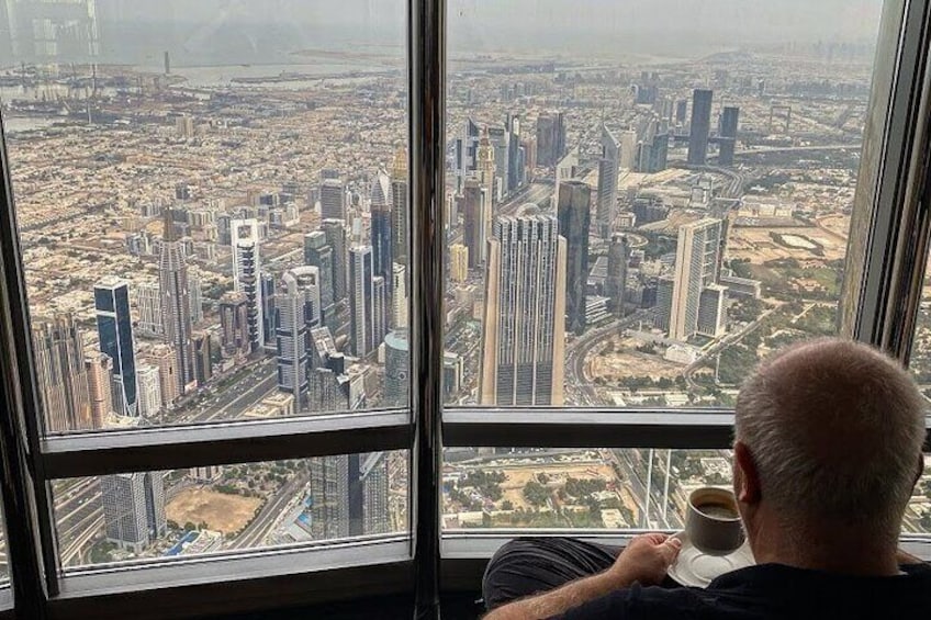 A man holding a cup, looking out of a large window at a cityscape with tall buildings and roads. 5