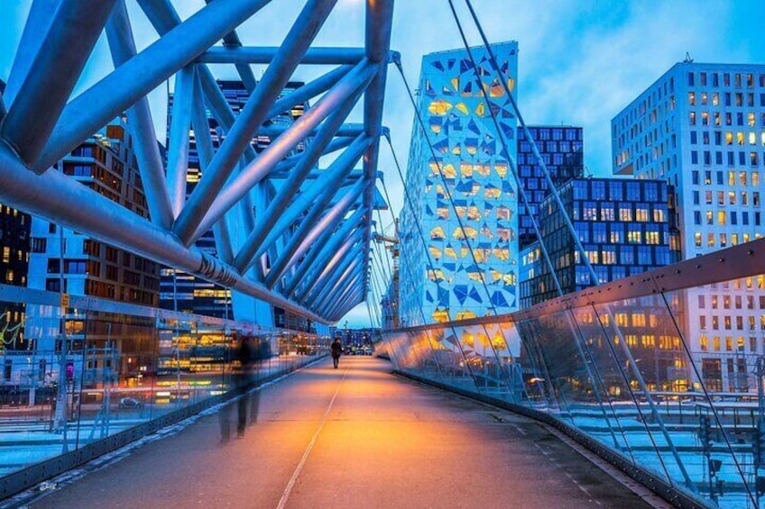 A pedestrian bridge with a metal framework and glass panels, overlooking a cityscape with modern buildings. 5