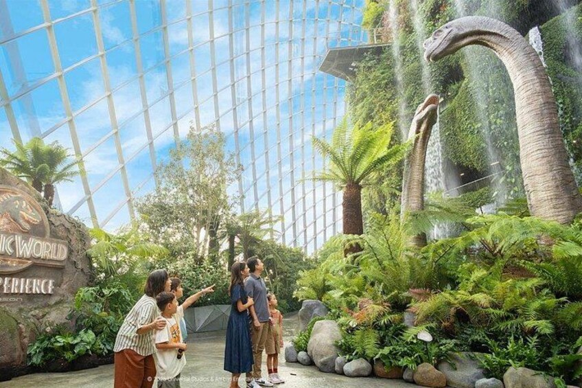 Un groupe de personnes se trouve dans un jardin intérieur luxuriant, doté d'un grand plafond en verre, d'une enseigne indiquant « The World Experience » et d'une grande sculpture de dinosaure. 2