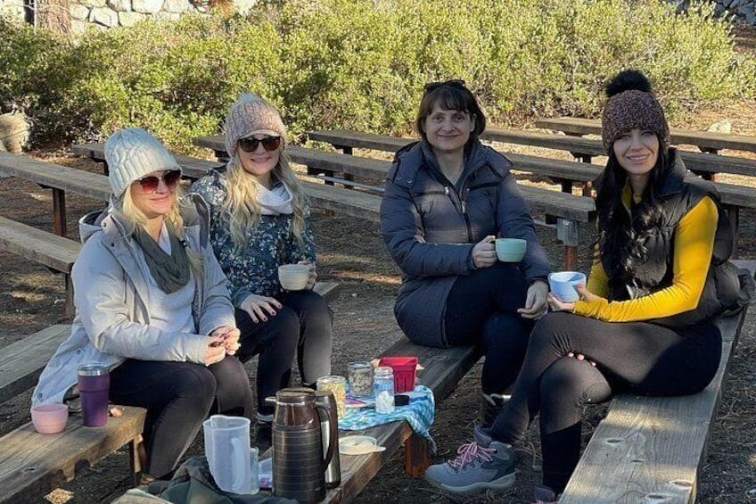 Quatre personnes sont assises sur des bancs en bois à l'extérieur, devant une table sur laquelle sont posées des tasses et un thermos. 5