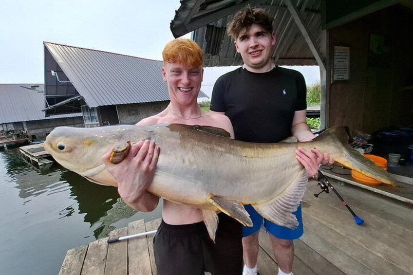 Two people holding a large fish on a dock with a boat and buildings in the background. 4