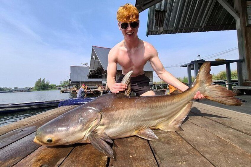 A person holding a large fish on a wooden dock with a boat and a building in the background. 5