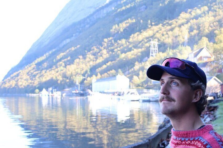 Una persona de pie junto a un lago tranquilo con barcos, un faro y casas en una ladera. 3