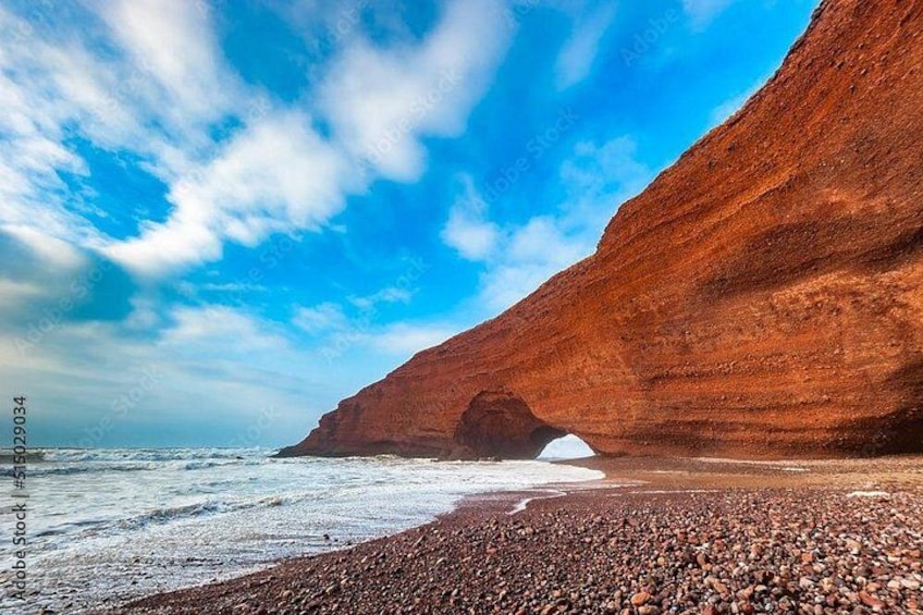 A coastal landscape with a prominent red cliff, a pebbly beach, and a clear blue sky. 1