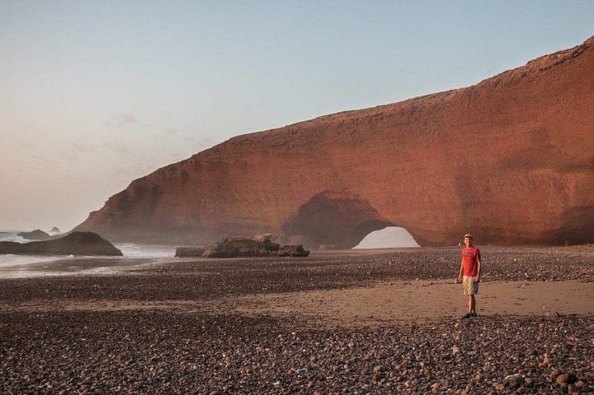 A person standing on a pebbly beach with a large red rock formation in the background. 3