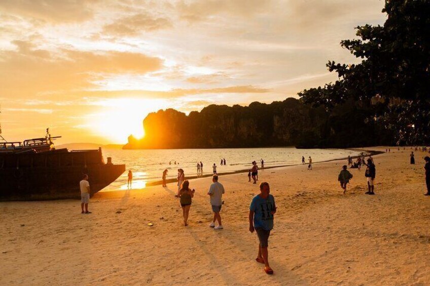 A beach at sunset with people walking and standing on the sand, a boat docked on the left, and a rocky cliff in the background. 2