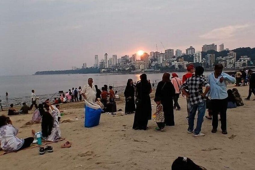 Una playa concurrida con gente sentada y de pie, un atardecer al fondo y el horizonte de una ciudad al otro lado del agua. 5