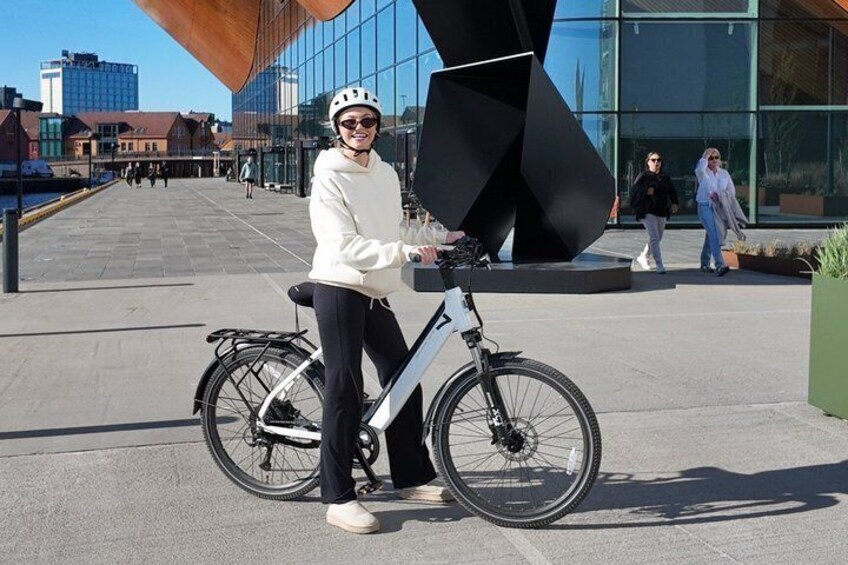 A person standing next to a bicycle in an urban setting with modern architecture and a waterfront in the background. 5