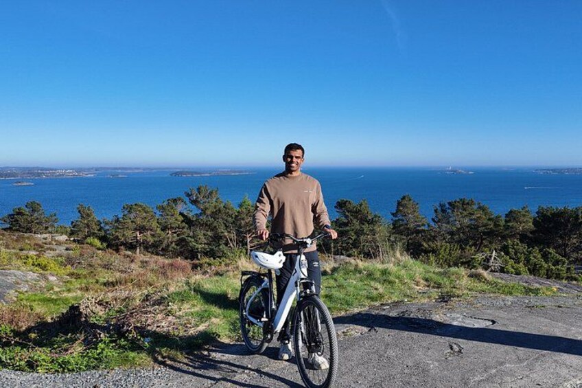 A person standing next to a bicycle on a coastal road with a view of the ocean and distant land. 4