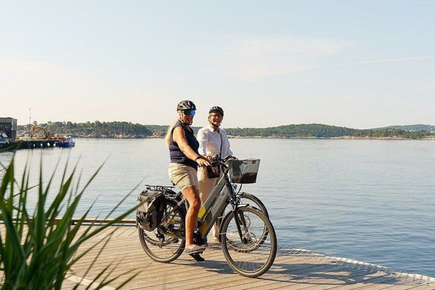 Two people on a stationary bicycle by a calm body of water with a distant shoreline. 1