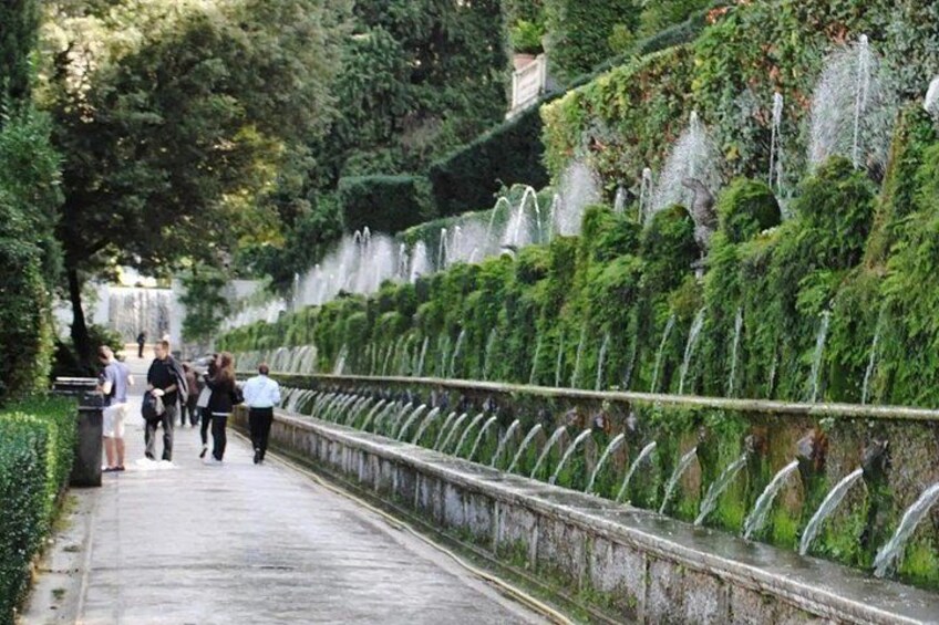 A pathway lined with neatly trimmed hedges and a series of water fountains. 2