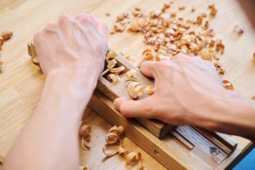 Des mains utilisent un rabot sur une surface en bois, et des copeaux de bois sont éparpillés autour. 2