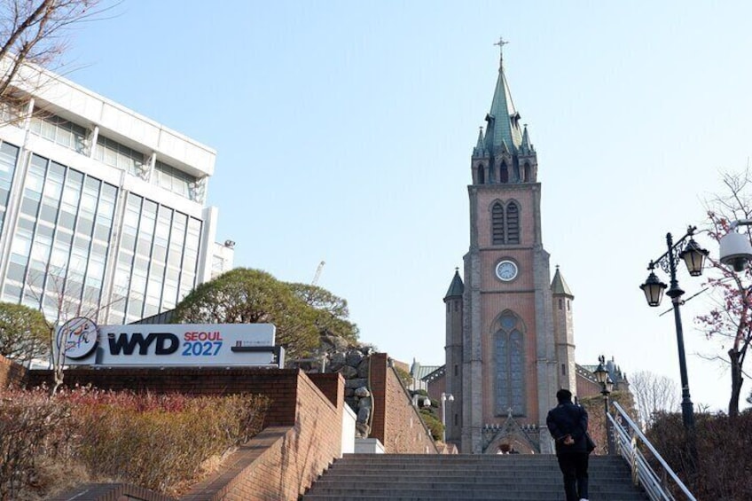 Une église dotée d’un haut clocher, une pancarte indiquant « JMJ 2027 », et une personne qui monte les escaliers. 2