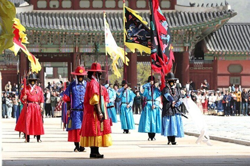 Une cérémonie traditionnelle coréenne, où les participants, vêtus d’hanboks aux couleurs vives, brandissent des drapeaux et se produisent devant un bâtiment historique. 1