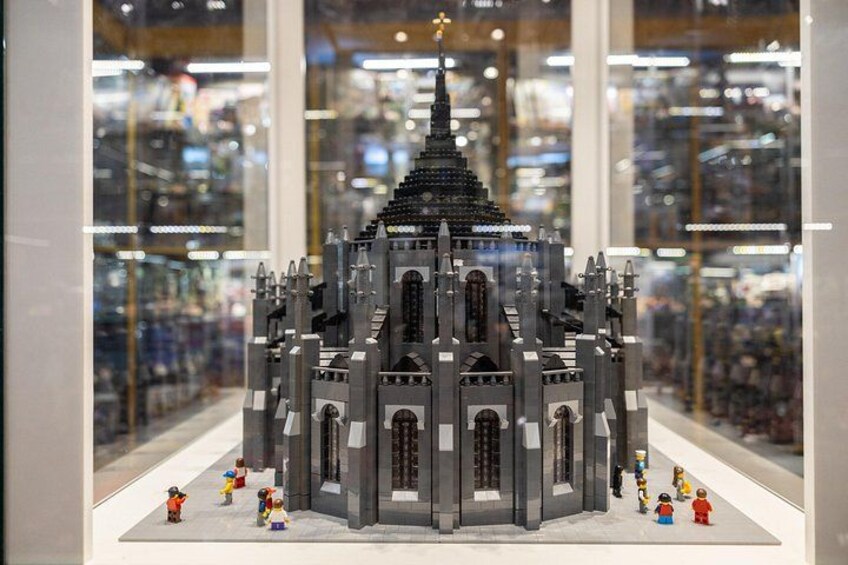 A detailed LEGO model of a gothic-style building with multiple towers and a spire, displayed in a glass case with miniature LEGO figures around it. 1