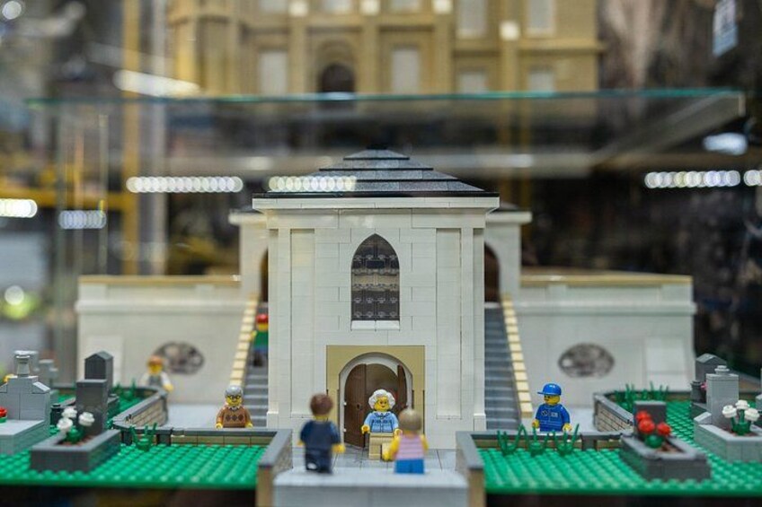 A LEGO model of a building with a central archway, two figures standing in front, and a garden area with plants and flowers. 5