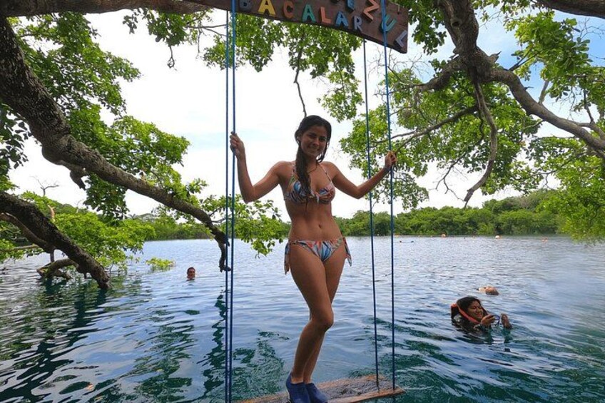 Una persona con traje de baño de pie sobre una plataforma flotante en un cuerpo de agua, rodeada de árboles y un letrero que dice 'BACALAO'. 2