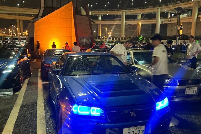 A nighttime urban scene with a blue-lit car in traffic, a large orange structure, and a busy street with pedestrians. 4