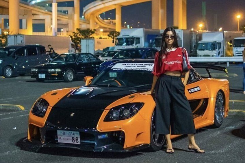 A woman standing next to a bright orange sports car in a parking lot at night. 1