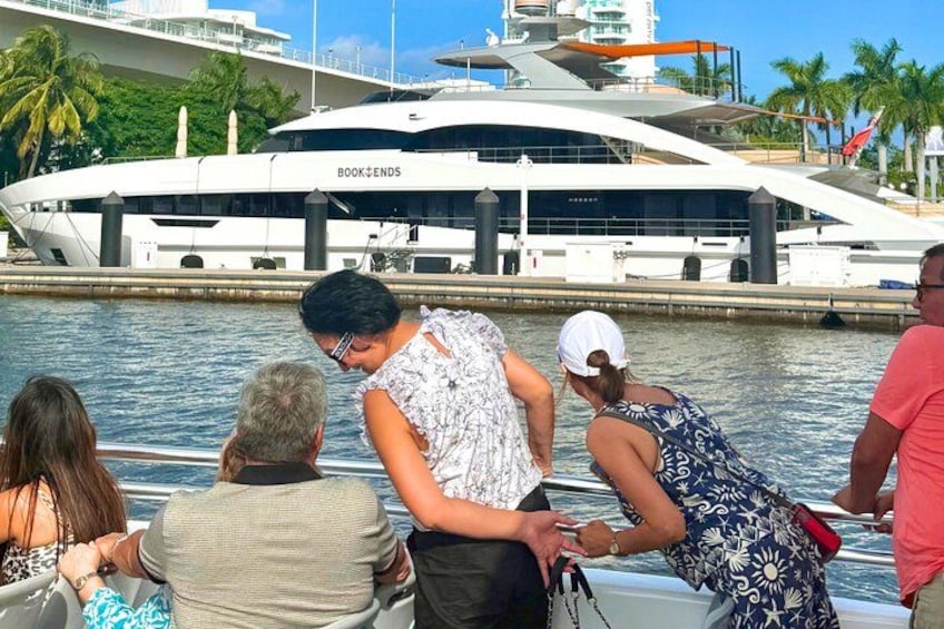 Un groupe de personnes se trouve sur un bateau près d’un grand yacht baptisé « BOOKENDS », qui est amarré dans une marina, avec des palmiers en arrière-plan. 2