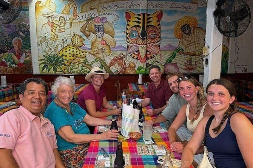 A group of people seated around a table with a colorful tablecloth, a bottle, and a plate of food. 1