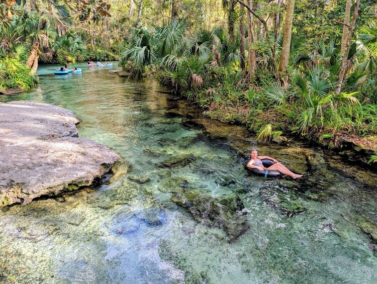 A clear stream with people floating in tubes, surrounded by lush greenery and rocks. 1
