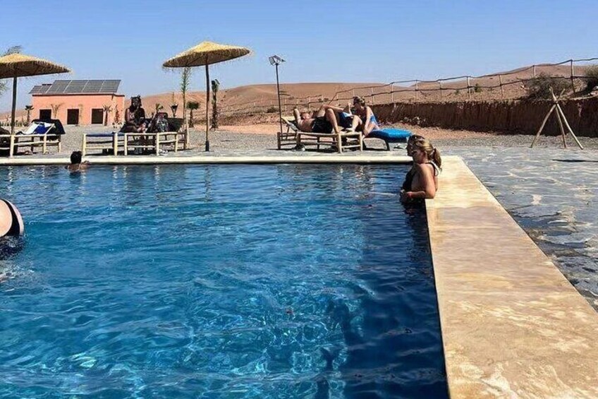 En swimmingpool med folk, der slapper af under parasoller, en bygning i baggrunden og et ørkenlandskab. 1