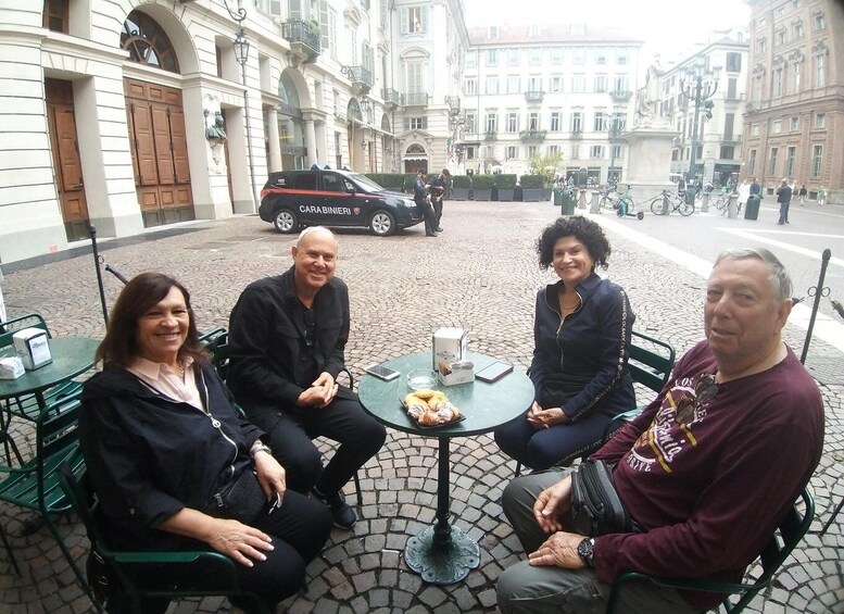 Four people sitting at a table with a croissant and a glass of water. A police car is parked in the background. 2