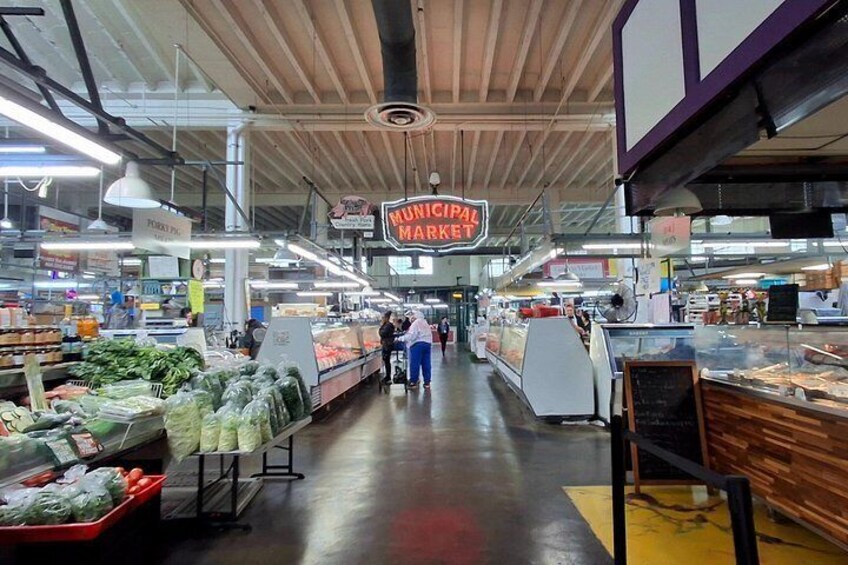 Een levendige overdekte markt met verschillende afdelingen vol groenten en fruit, en een opvallend bord waarop 'Stadsmarkt' staat. 2