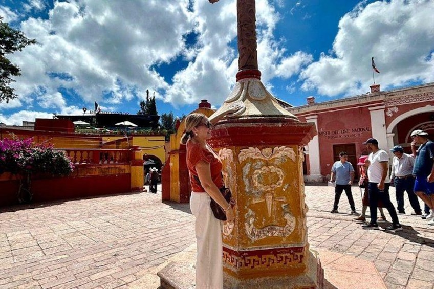 Eine Person steht in der Nähe einer verzierten Säule mit Inschriften, einem Gebäude mit roter Fassade und einem klaren, blauen Himmel mit Wolken. 4