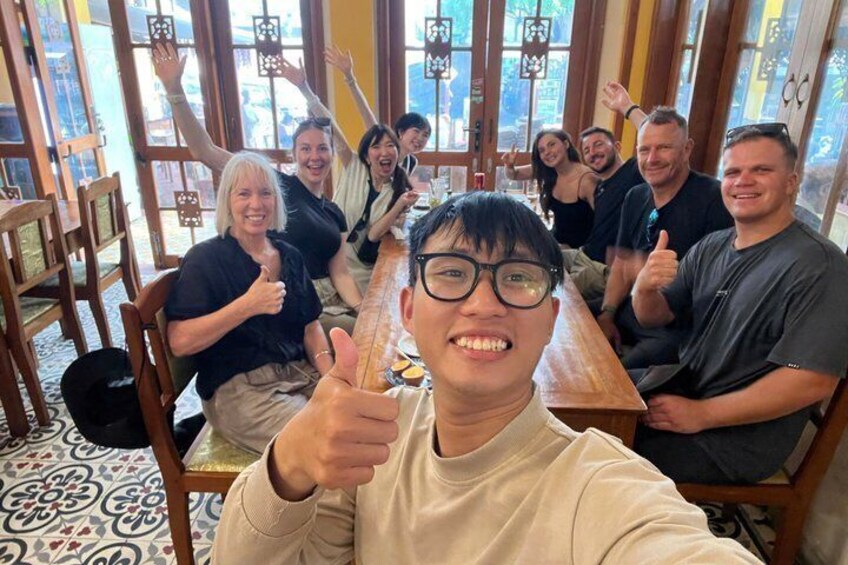 A group of people sitting at a wooden table in a restaurant with patterned floor tiles and large windows. 2