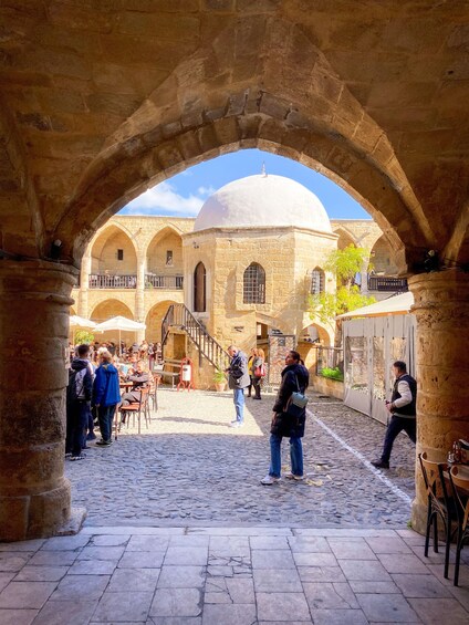 An ancient stone courtyard with arches, a dome, and people walking. 1