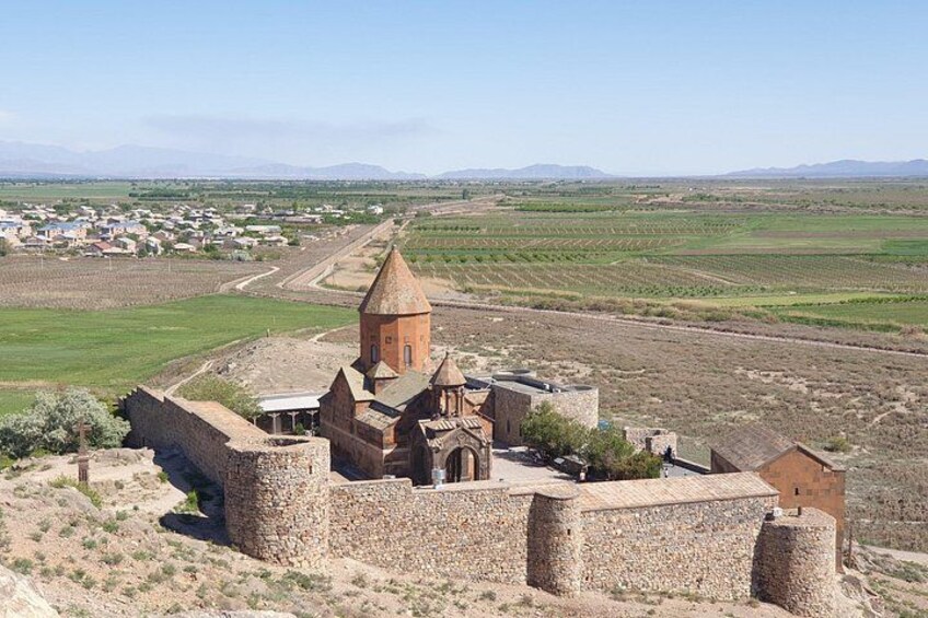 A historical stone fortress with a church, surrounded by fields and distant mountains. 1
