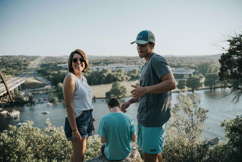 Three people standing by a river with a bridge and buildings in the background. 3