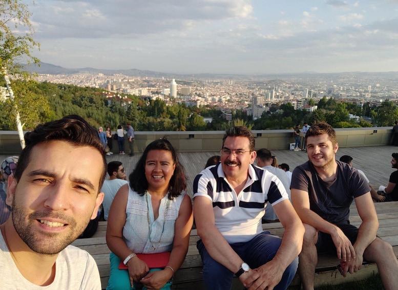 A group of people sitting on a wooden deck overlooking a cityscape with mountains in the background. 1
