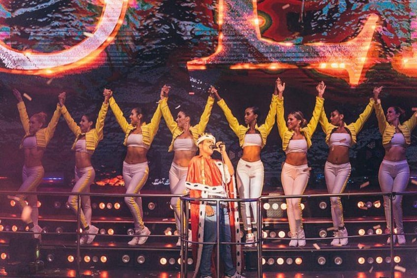 Un grupo de personas en un escenario con los brazos levantados, vistiendo chaquetas amarillas y pantalones blancos, con un letrero iluminado grande al fondo. 3